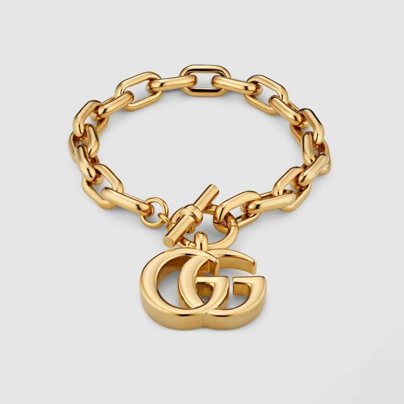 Gold chain bracelet with a heart-shaped Gucci logo charm on a light gray background