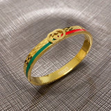 A gold-tone Gucci bangle bracelet with green and red enamel stripes and a double G logo, resting on a textured gray fabric background.