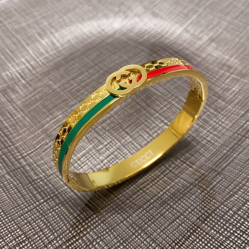 A gold-tone Gucci bangle bracelet with green and red enamel stripes and a double G logo, resting on a textured gray fabric background.